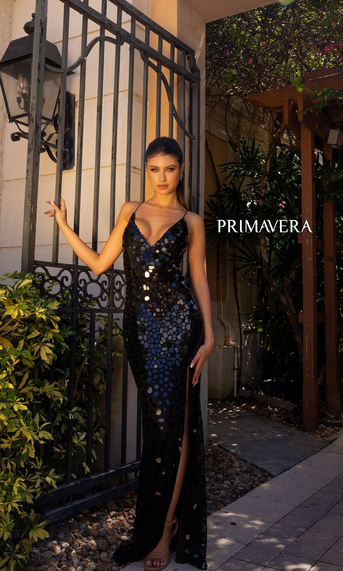 A woman stands by a wrought iron gate in lush greenery, wearing the Primavera Broken-Glass Long Formal Dress 4151 with black sequins and a thigh-high slit;